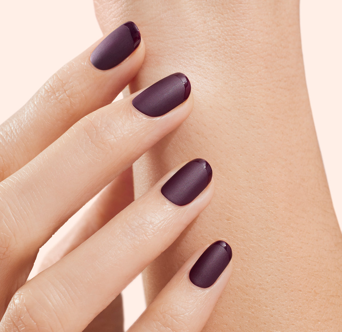Gros plan d'une main aux ongles courts et soigneusement manucurés, vernis d'une couleur prune foncé mat, touchant délicatement le poignet de l'autre main sur fond clair.