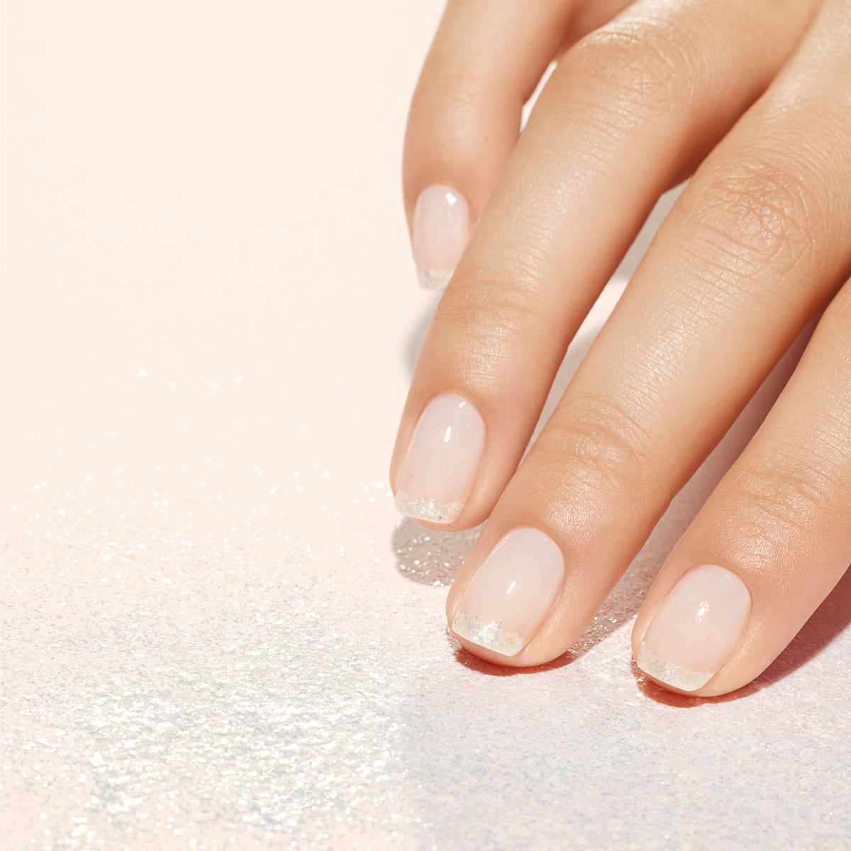 A hand with neatly manicured nails featuring a nude-pink polish and subtle glittery tips, resting on a light, sparkly surface.