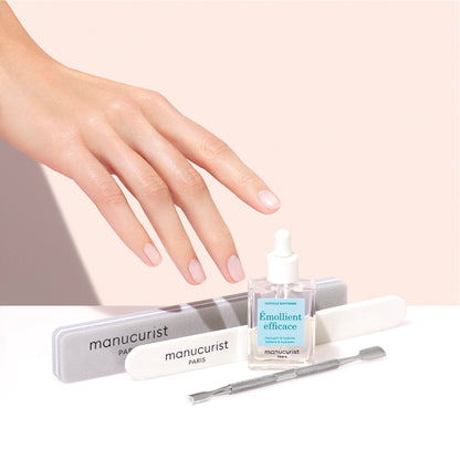 A hand with manicured nails hovers over Manucurist nail care essentials, including the Hydrating Cuticle Softener, emollient oil, nail files, and a cuticle pusher—all set against a soft pink background.