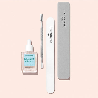 A bottle of Manucurist Hydrating Cuticle Softener, alongside a branded metal cuticle pusher, white nail file, and gray nail buffer, are arranged neatly on a light pink background—creating the perfect set for an at-home manicure.
