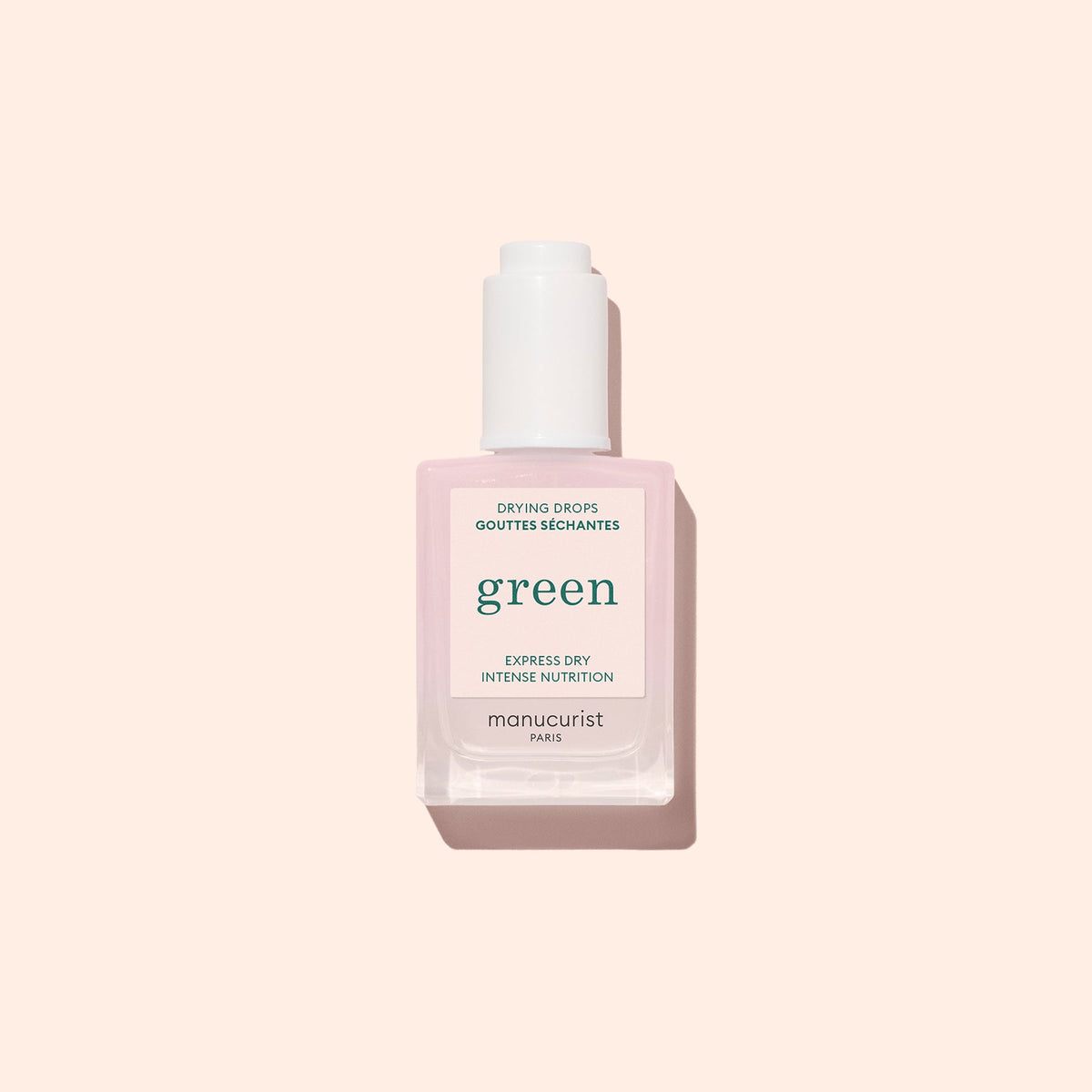 A bottle of Manucurist Paris green Drying Drops for nails, with a light pink label and a white cap, is placed on a solid pale pink background.