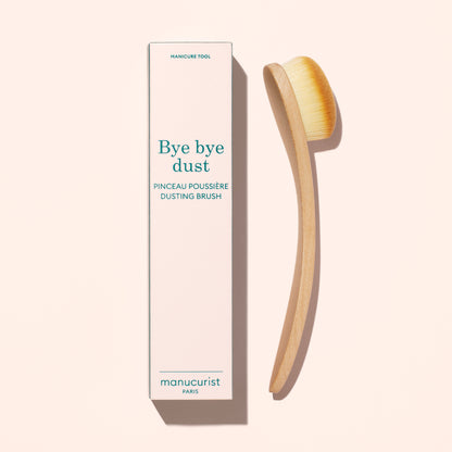 A Dust Brush by Manucurist with vegan bristles and a curved wooden handle sits beside its tall box labeled “Bye bye dust – Dust Brush” on a light beige background.