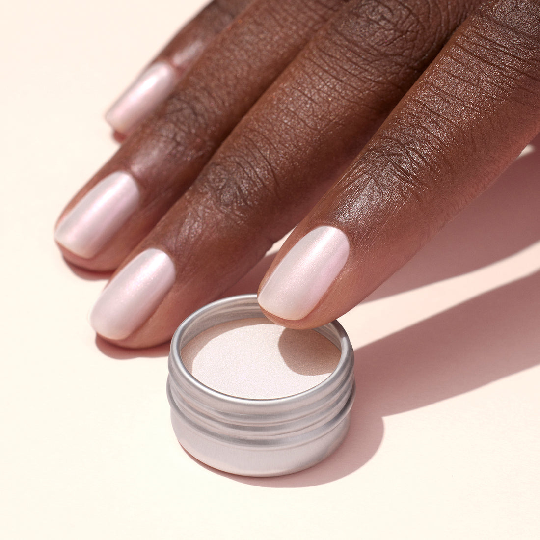 A hand with neatly manicured, pale pink pearlescent nails is shown beside an open small silver container of Manucurist Pink Glazed Effect Powder on a light background.