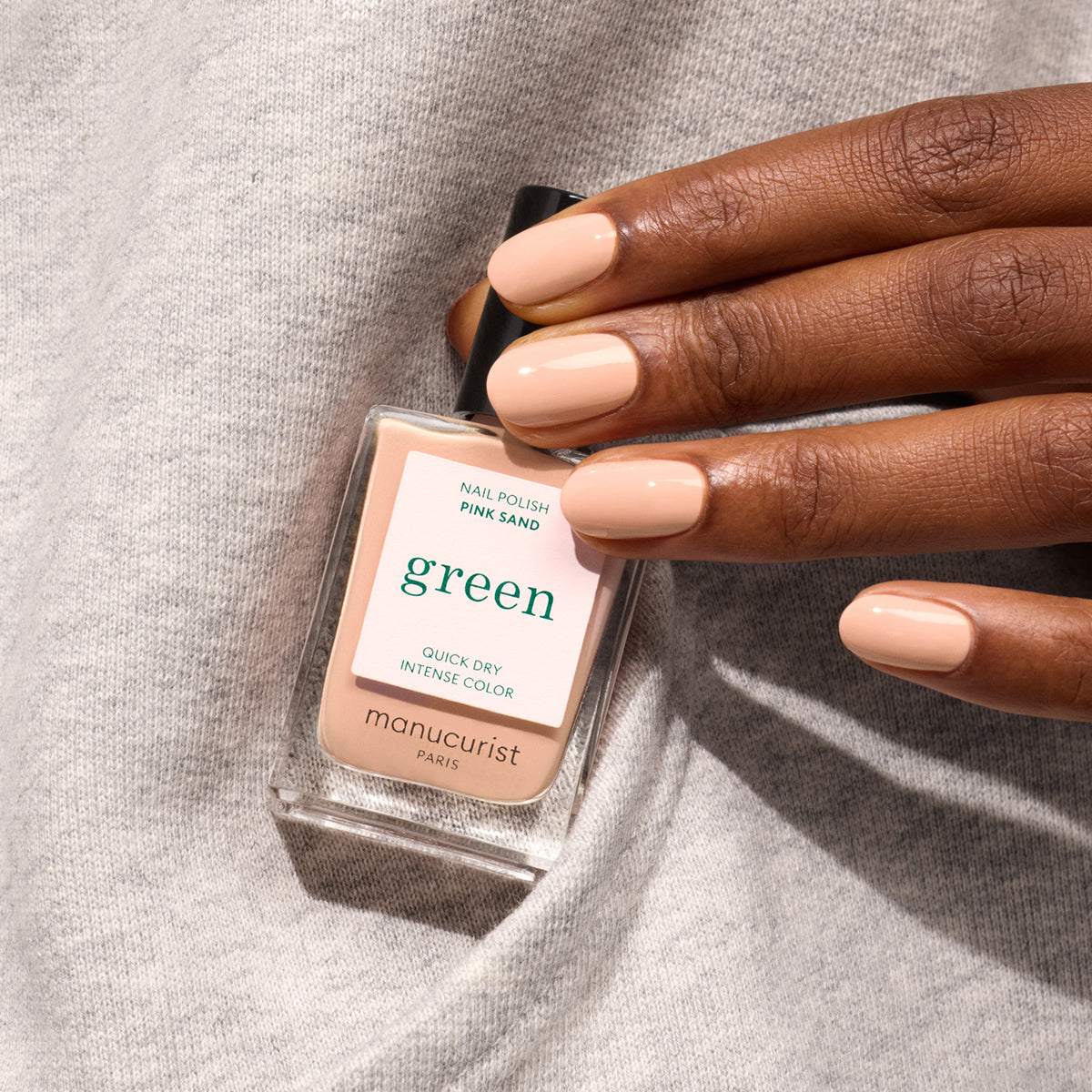 A hand with neatly manicured nails painted in Pink Sand, a pale nude shade by Manucurist, holds a matching bottle of nail polish against soft gray fabric.