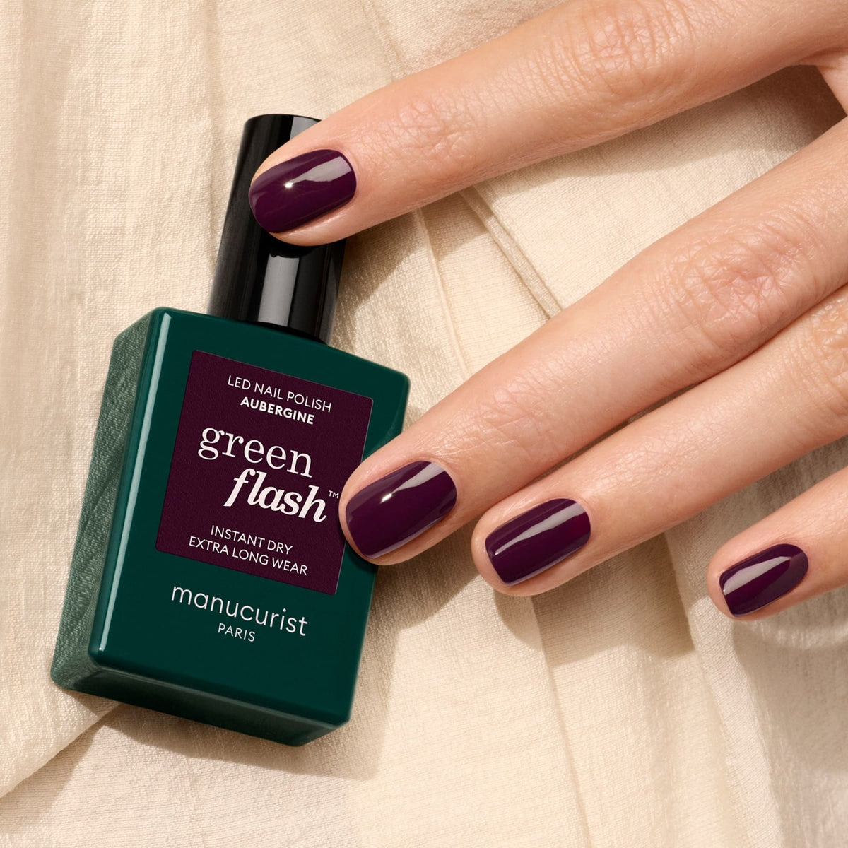A hand with neatly manicured nails painted in the dark Aubergine shade holds a green bottle of Manucurist&
