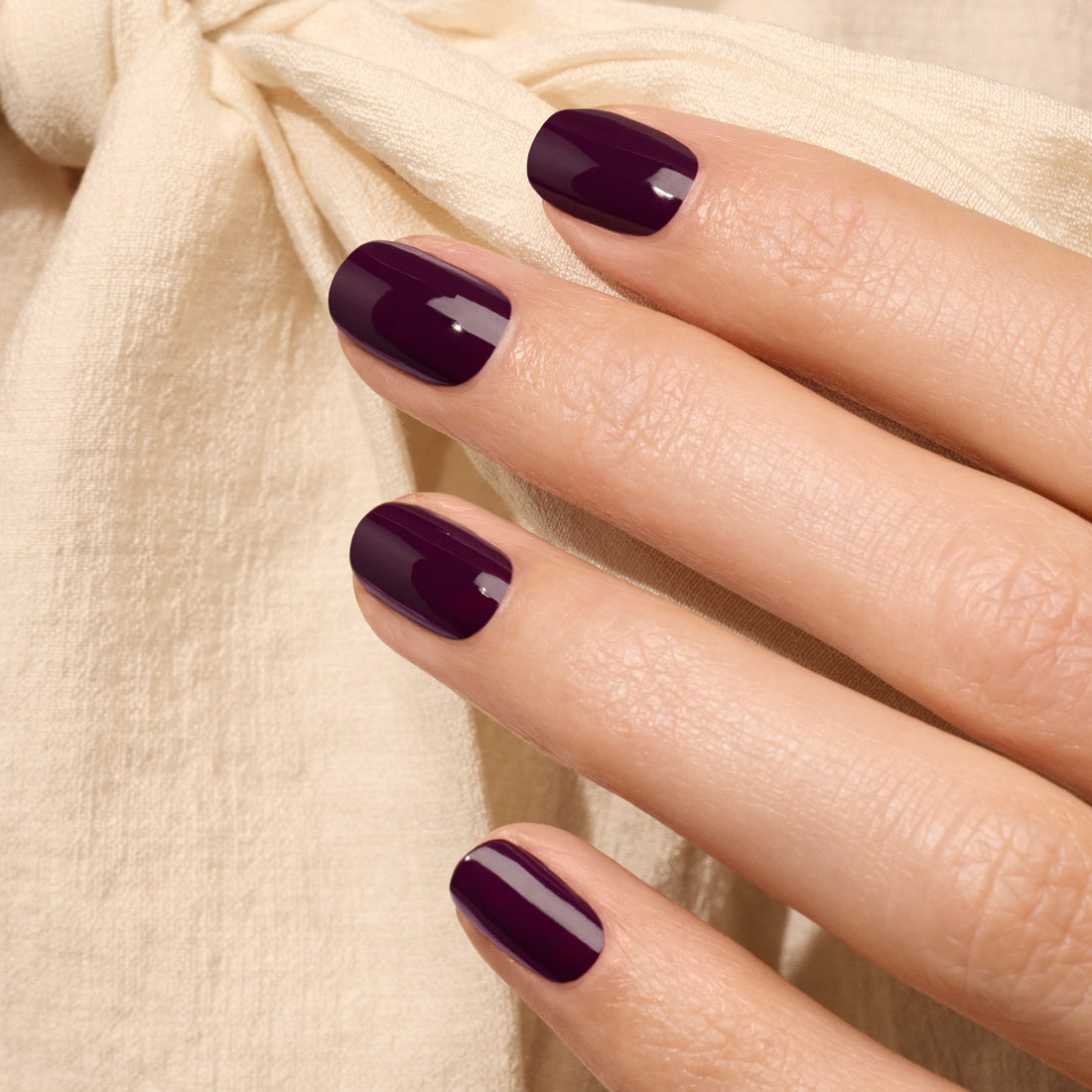 Une main aux ongles soigneusement manucurés, vernis en Aubergine par Manucurist, repose sur un tissu beige clair orné d&