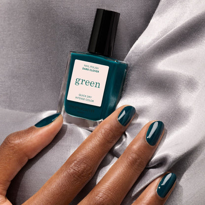 Une main aux ongles vert sarcelle foncé tient un flacon de vernis à ongles Manucurist Dark Clover, posée sur un tissu en satin gris.