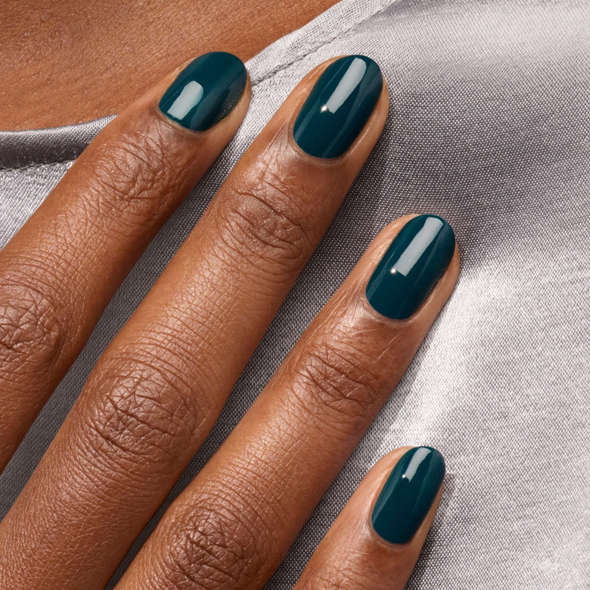 Gros plan d’une main aux ongles soigneusement manucurés, vernis du vernis bleu-vert brillant "Dark Clover" de Manucurist, reposant sur un tissu en satin gris clair.