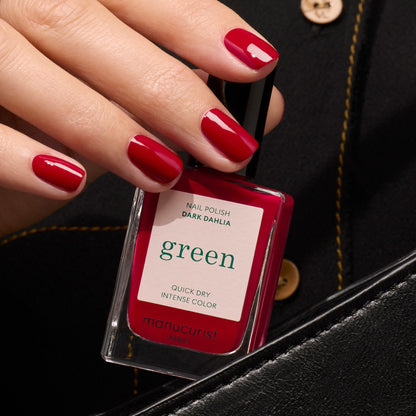 Une main aux ongles rouges brillants tient un vernis à ongles Manucurist Dark Dahlia — une teinte rouge foncé intense — devant une tenue noire à boutons dorés.