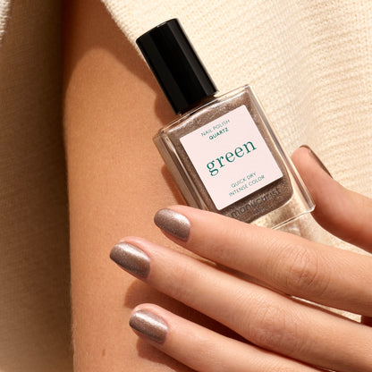 A hand with taupe nail polish holds a square Manucurist bottle labeled "Quartz" with a black cap, set against a beige fabric background.