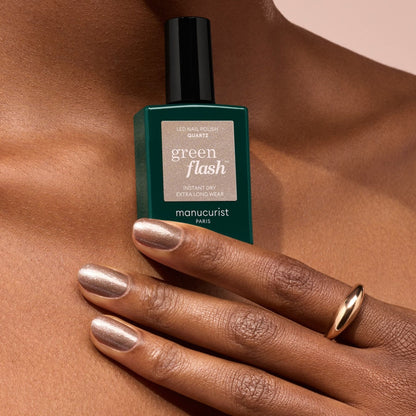 A hand with chestnut brown, semi-permanent polished nails holds a green bottle of Manucurist Quartz nail polish. The person wears a simple gold ring, and the background is soft beige.