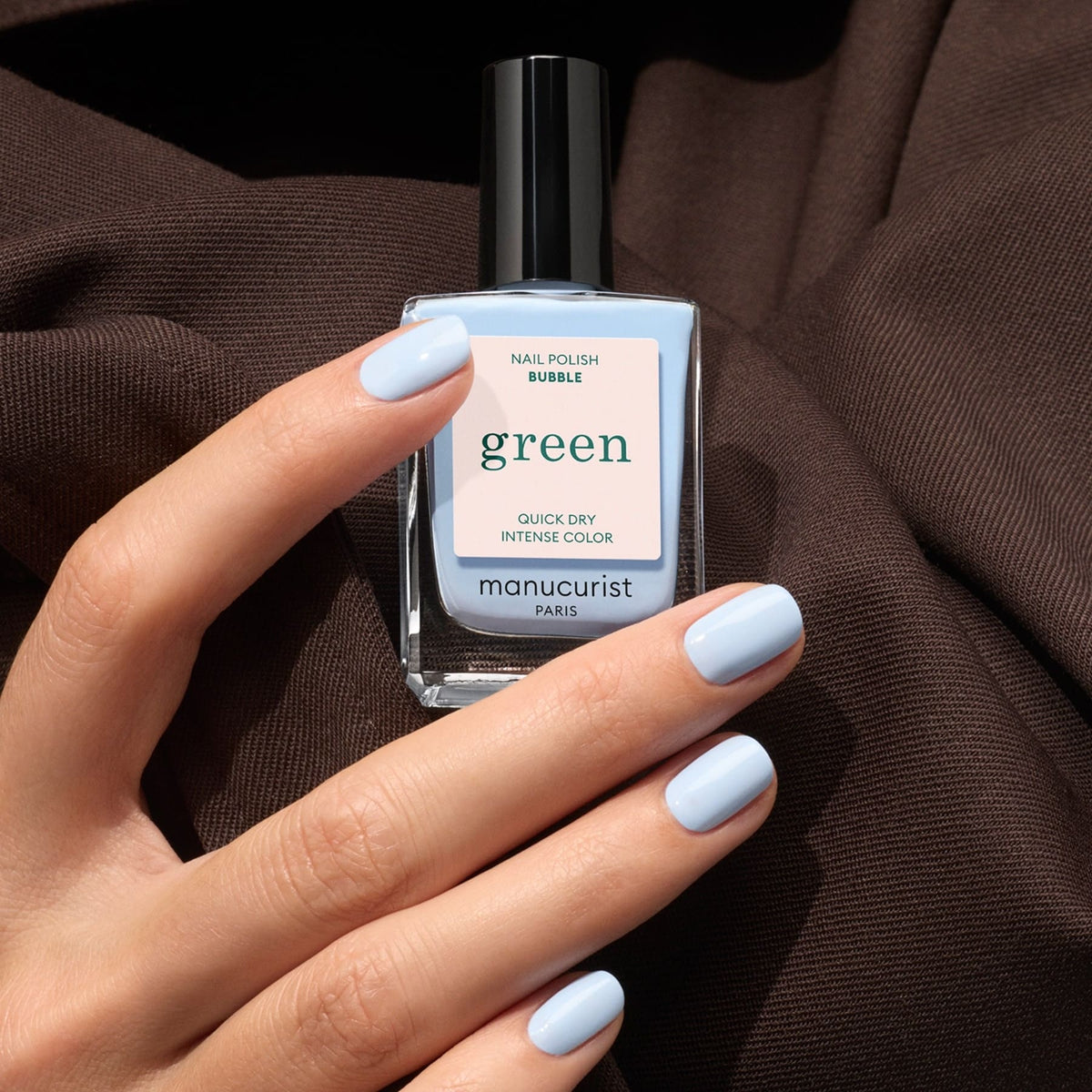 A hand with light blue painted nails holds a bottle of Manucurist Bubble nail polish, a pastel blue shade with grey undertones, against a brown fabric background.