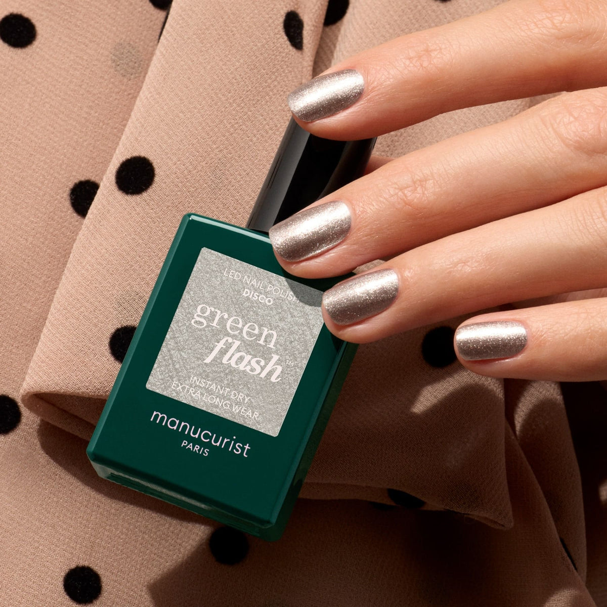 A hand with opaque glittery silver nail polish in the shade Disco by Manucurist holds a green bottle of gel polish against beige fabric with black polka dots.