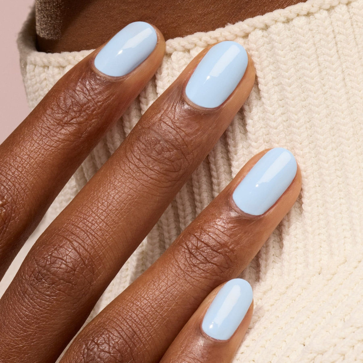A hand with medium-brown skin rests on a cream knit sweater, displaying neatly manicured nails painted in Manucurist’s Bubble, a smooth, glossy light blue nail polish with subtle grey undertones.