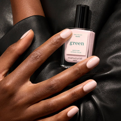 A hand with neatly manicured iridescent pink nails holds a bottle of Manucurist Gloss nail polish against a black leather background.