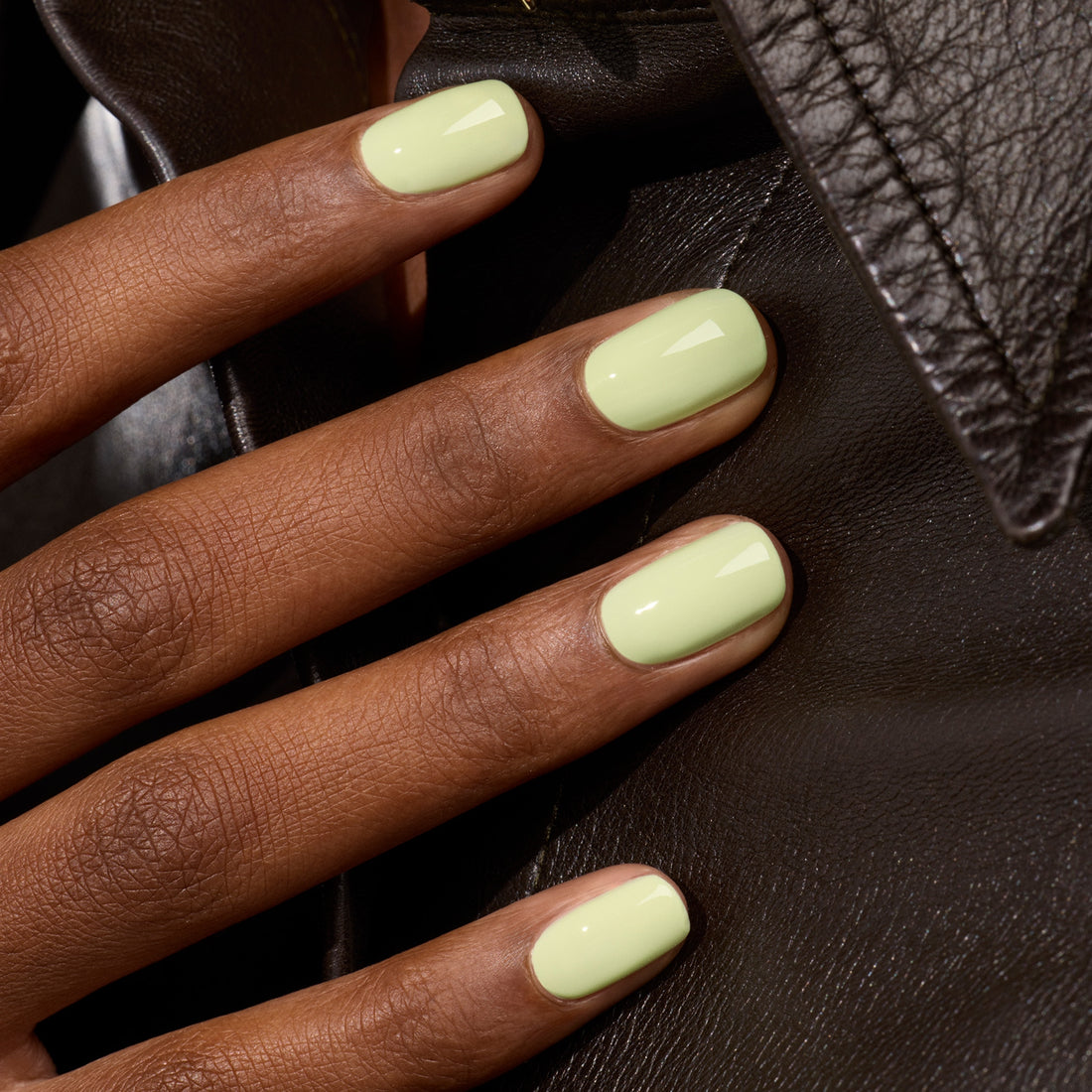 A hand with short, neatly filed nails painted in Pistachio, a glossy pastel yellow shade from Manucurist’s Green™ range, rests on a black leather jacket.
