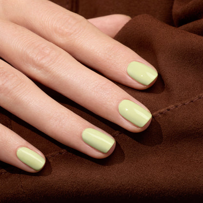 Gros plan sur une main aux ongles soigneusement manucurés, peints avec Pistachio de Manucurist, un vernis gel LED vert pâle aux doux sous-tons jaunes, reposant sur une surface en tissu marron.