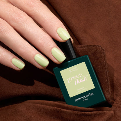 Une main aux ongles soigneusement manucurés, peints avec Manucurist Pistachio, un vernis gel LED vert pâle aux sous-tons jaunes, tient le flacon sur un fond en tissu marron.