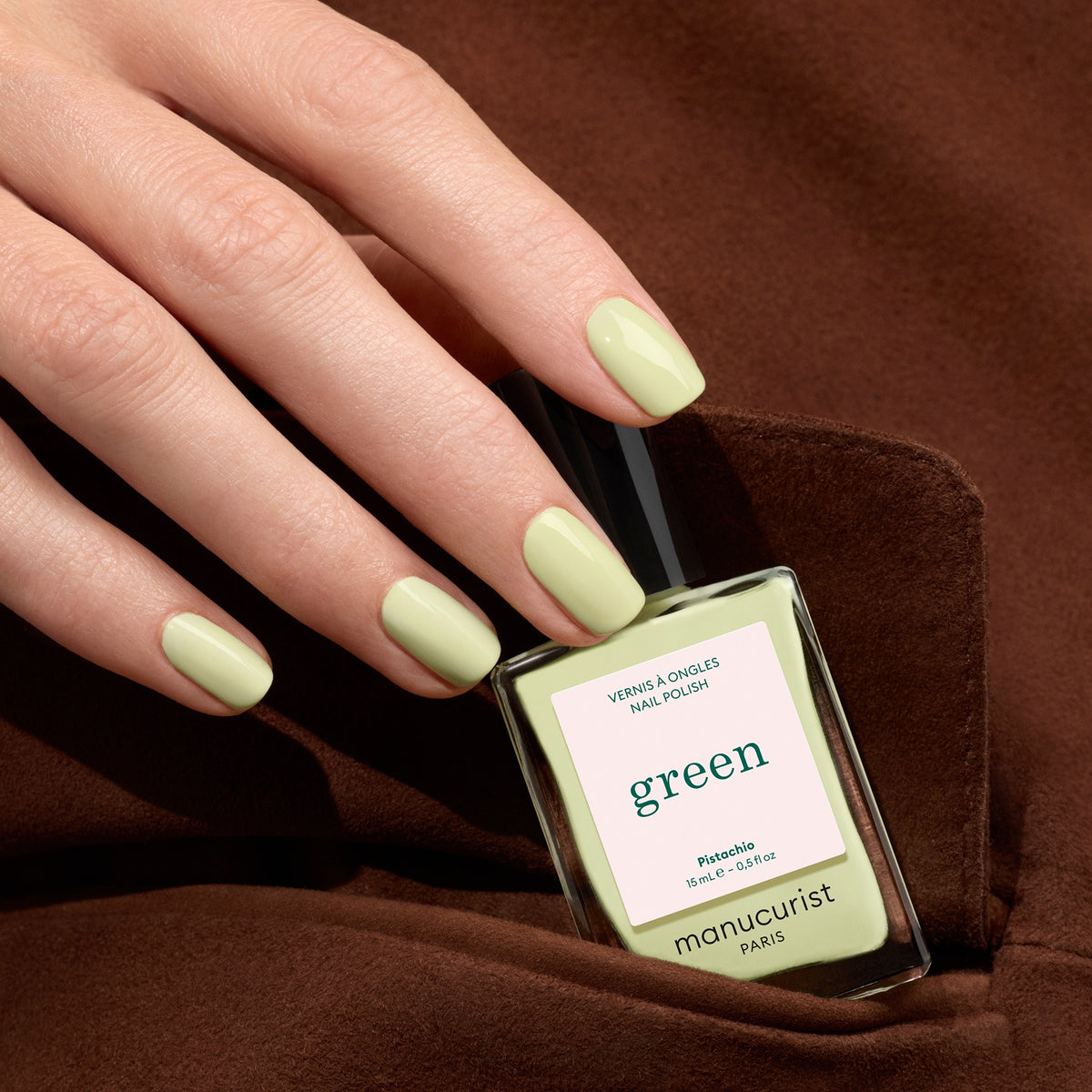 A hand with neatly manicured nails painted in Pistachio by Manucurist holds the matching bottle, showing off its pale green shade with yellow undertones against a brown fabric background.