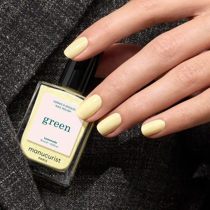 A hand with neatly manicured nails painted pale yellow holds a matching bottle of Lemonade by Manucurist, set against a textured dark fabric background.