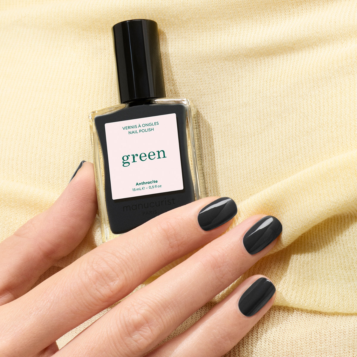 A hand with neatly manicured, glossy dark grey nails holds a bottle of Manucurist Anthracite nail polish against a soft yellow fabric background.