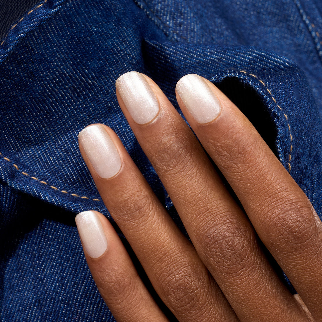 Une main à la peau brun moyen et aux ongles soigneusement manucurés, peints avec Moon de Manucurist, un vernis blanc nacré, repose sur un tissu en denim bleu aux surpiqûres visibles.