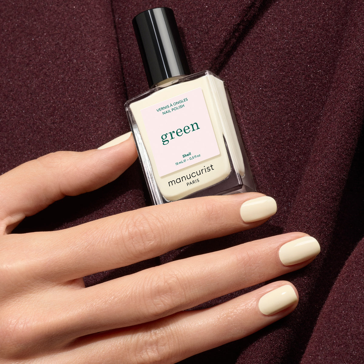A hand with off-white, neatly manicured nails holds a bottle of Manucurist Shell nail polish against a maroon fabric background.