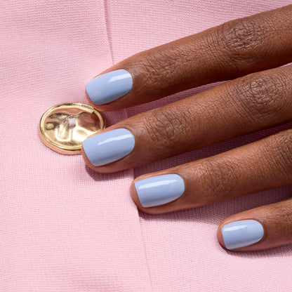 Une main aux ongles soigneusement manucurés, vernis dans la teinte Lilas mauve brillante de Manucurist, posée sur un tissu rose avec un bouton doré.