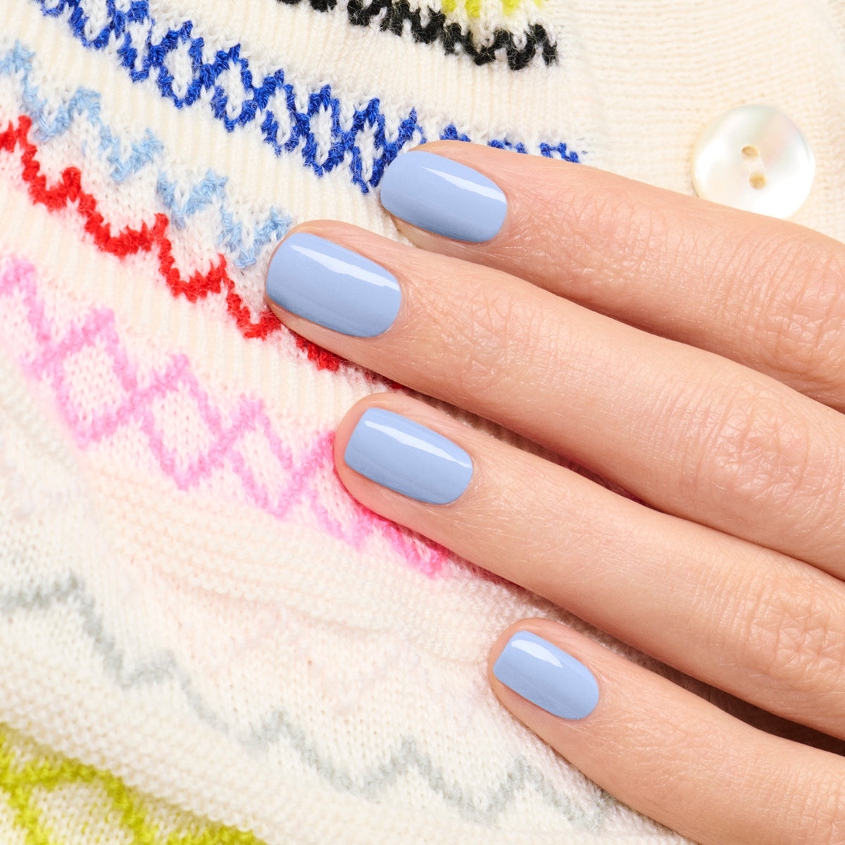 Une main aux ongles courts et soigneusement manucurés, peints avec le vernis à ongles bleu pastel Lilas de Manucurist, repose sur un pull blanc en maille orné de motifs zigzag colorés et d’un bouton blanc.