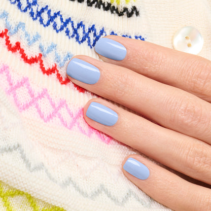 Une main aux ongles courts et soigneusement manucurés, peints avec le vernis à ongles bleu pastel Lilas de Manucurist, repose sur un pull blanc en maille orné de motifs zigzag colorés et d’un bouton blanc.
