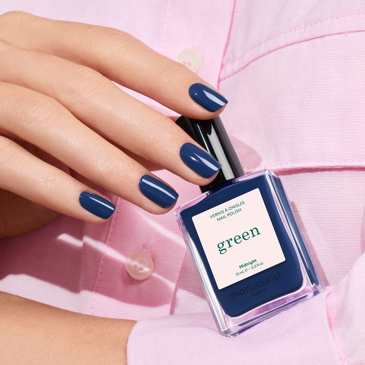 Une main aux ongles impeccablement manucurés avec la teinte Midnight tient un flacon Manucurist assorti, sur fond de chemise rose pâle.