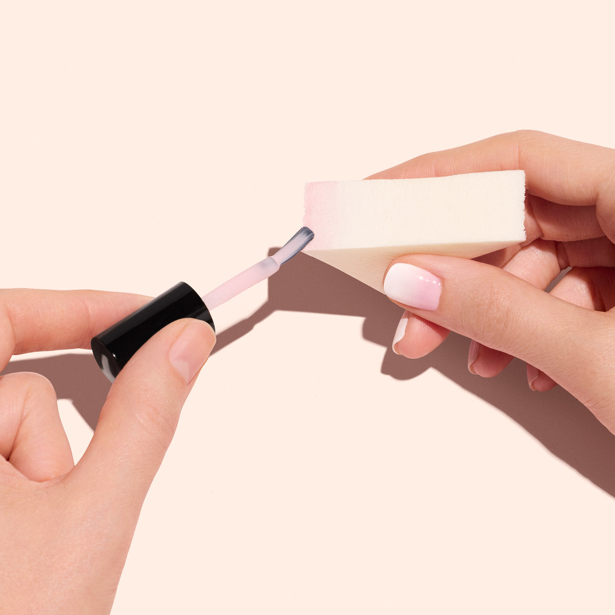 Une personne tient une Nail Art Sponge Manucurist (x4) dans une main et y applique du vernis rose clair à l’aide d’un pinceau. Ses ongles Baby Boomer soigneusement manucurés sont présentés sur un fond beige clair.