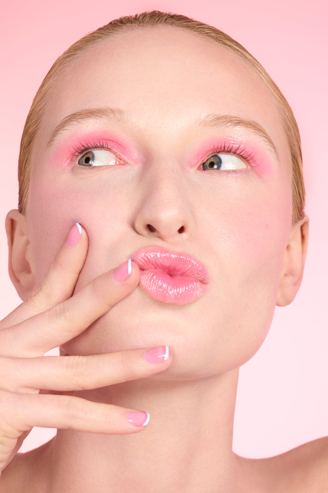 A person with slicked-back hair wears glossy pink lipstick, pink blush, and pink eyeshadow, touching their lips with manicured fingers against a soft pink background.