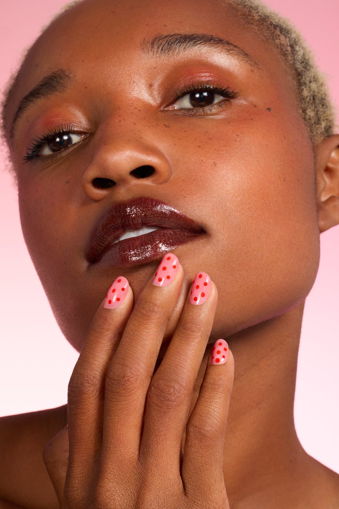 A person with short blond hair and glossy dark lips touches their face. Their nails are painted pink with red polka dots. The background is a soft pink gradient.