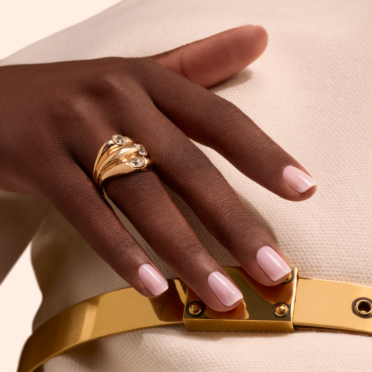 A hand with Manucurist Milky Pink nail polish rests on cream textured fabric, adorned with a large gold ring set with three gemstones. A shiny gold belt is also visible.