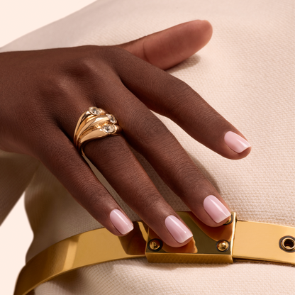 A hand with Manucurist Milky Pink nail polish rests on cream textured fabric, adorned with a large gold ring set with three gemstones. A shiny gold belt is also visible.