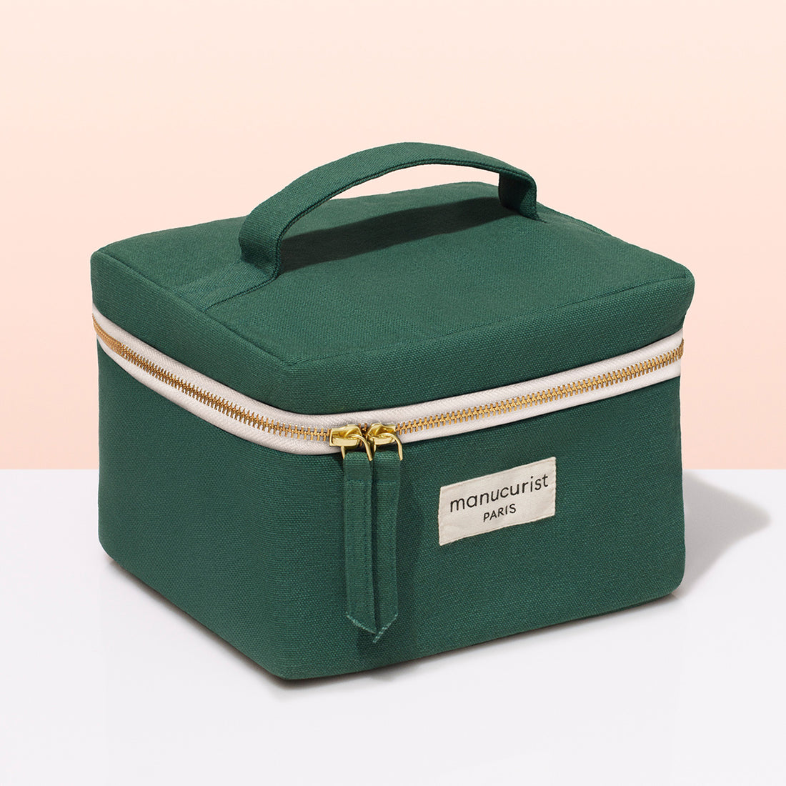 A green fabric Manucurist-test Vanity Case with a handle, gold zippers, and a beige "manucurist PARIS" label—ideal for nail polish or manicure storage—shown on a white surface with a light pink background.