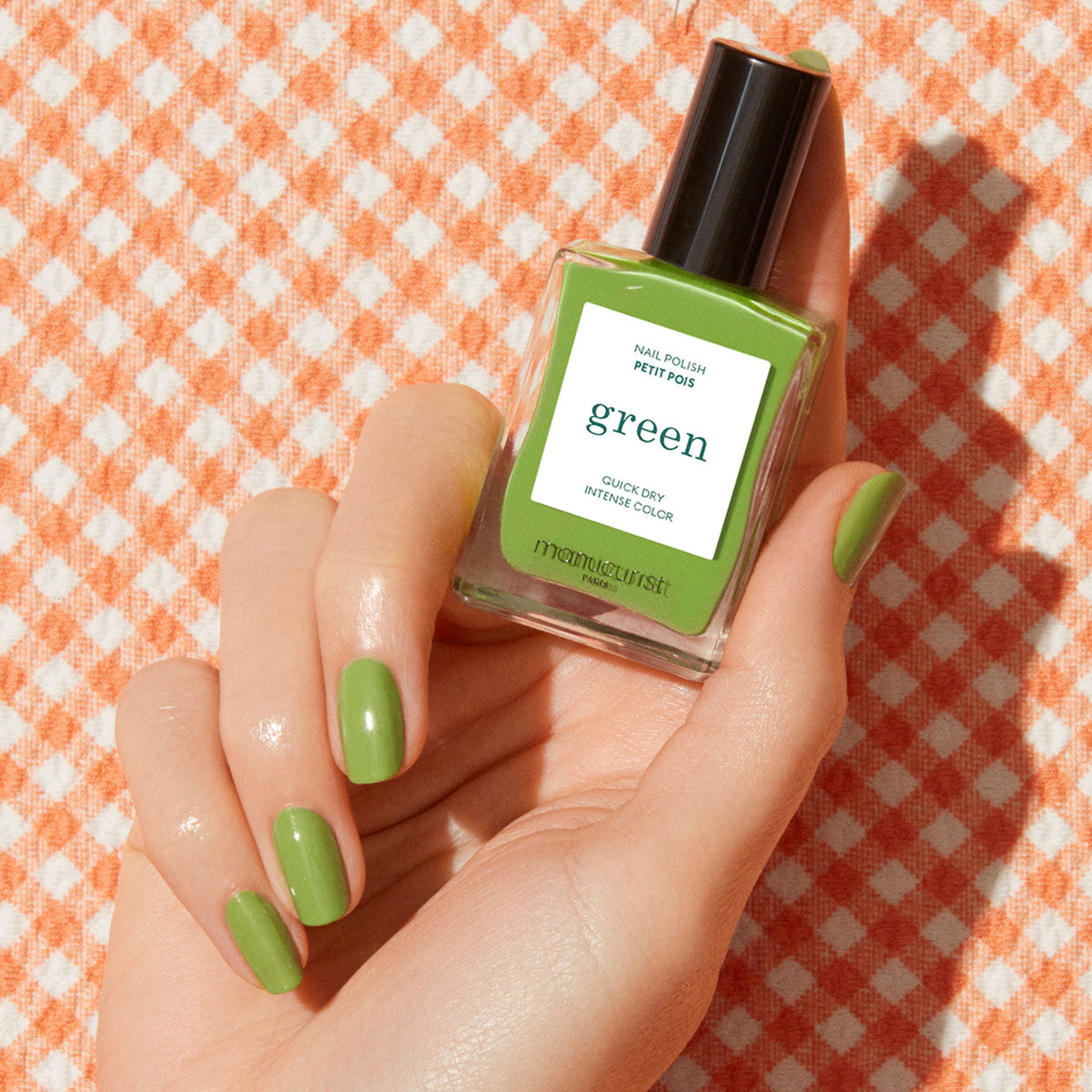 A hand with green painted nails holds a bottle of Manucurist Sweet Pea nail polish against an orange and white checkered fabric background.