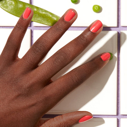 Une main à la peau foncée portant le vernis rose corail Pulp de Manucurist repose sur des carreaux blancs, entourée de pois gourmands et de petits pois de la gamme Green.