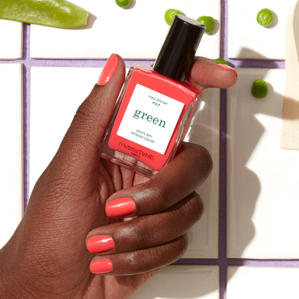 Une main à la peau mate affiche de jolis ongles corail tout en tenant un flacon de vernis Manucurist Pulp. En arrière-plan, des petits pois verts et une surface carrelée blanche aux joints lavande.