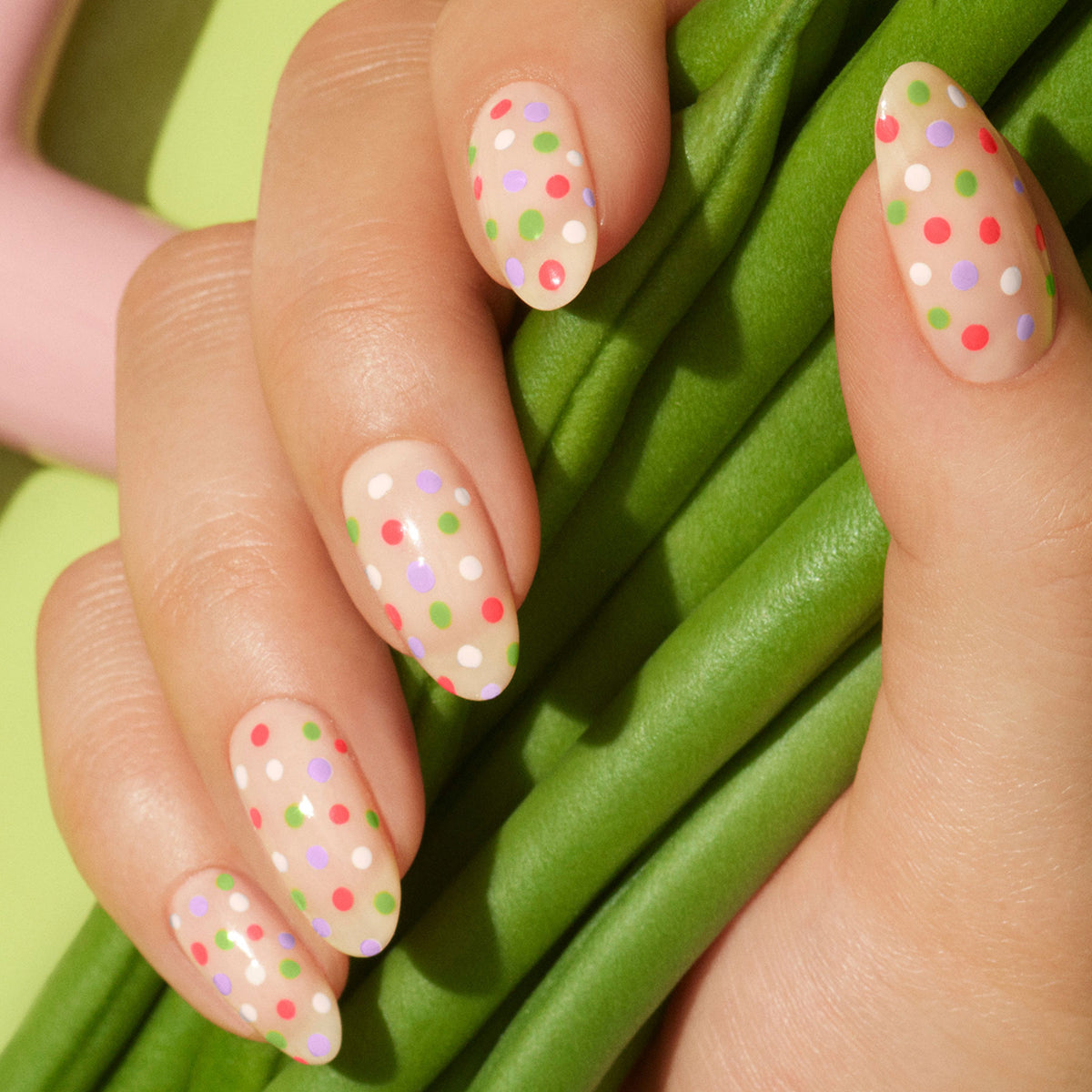 A hand with almond-shaped nails painted nude and decorated with colorful green, pink, white, and purple polka dots using Manucurist&