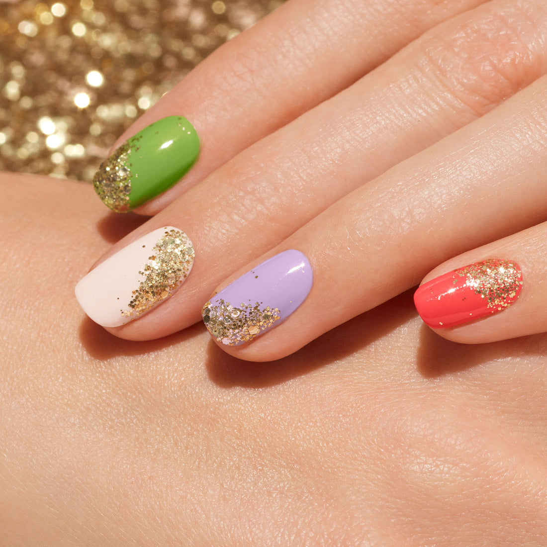 A hand with manicured nails in green, white, purple, and coral—each accented near the cuticles with Manucurist’s Glitter in Love biodegradable glitter—rests on another hand against a sparkling gold background.