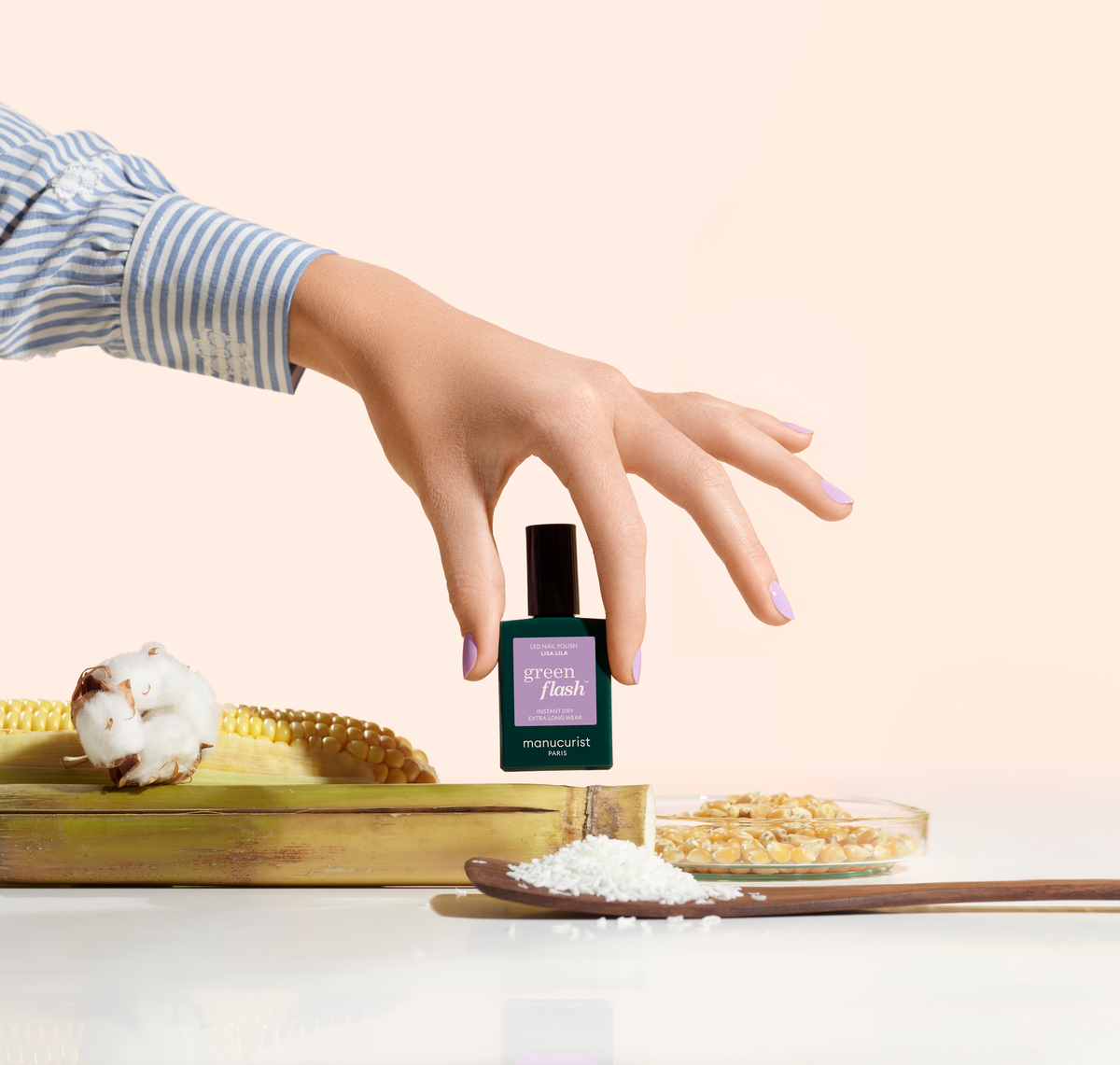 A hand with light purple nail polish holds a bottle labeled Green Flash above a corn cob, cotton, grains, and a wooden spoon with white granules, all arranged on a light surface.