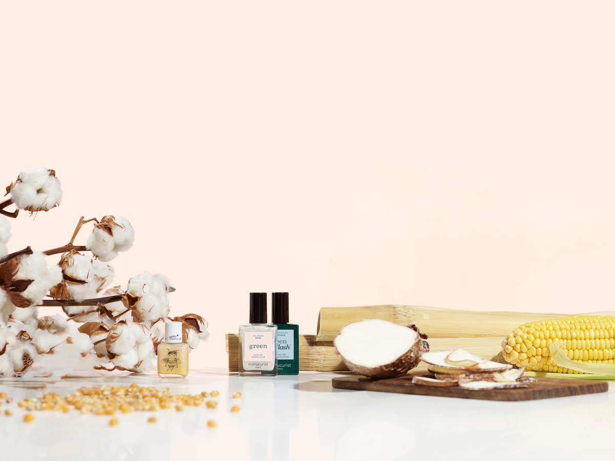 Bottles of nail polish displayed with cotton, bamboo, corn kernels, corn on the cob, and sliced root vegetables on a white surface with a light pink background.