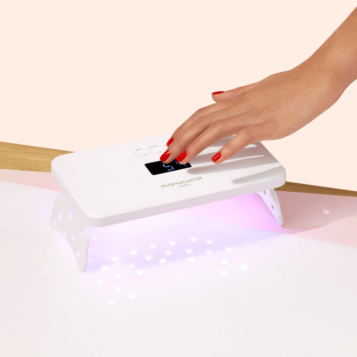 Une main avec un vernis Green Flash rouge catalyse sous la Lampe Manucurist Green Flash™ Premium 36W, qui émet une lumière violette pour un séchage rapide. La scène présente un arrière-plan clair et minimaliste.