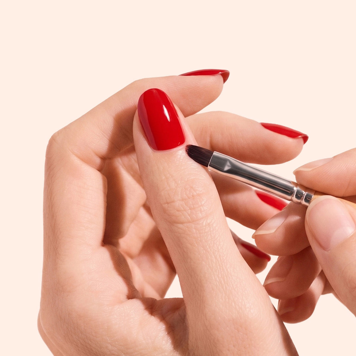 A hand with neatly manicured red nails uses a small brush to clean or perfect the edge of the nail polish. The background is a plain, light color.