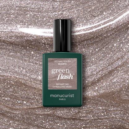 A bottle of Manucurist Quartz Green Flash semi-permanent nail polish sits on a glittery beige surface that matches the soft, refined pinkish-beige shade shown on the label.