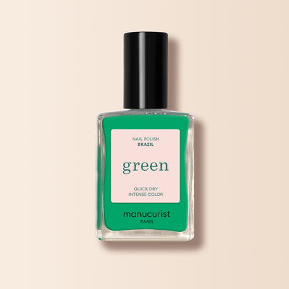 A clear glass bottle of Manucurist Brazil nail polish in electric green with a black cap. The label says Brazil, green, Quick Dry Intense Color, semi-matte finish by Manucurist Paris on a pink and green background. Perfect with your favorite Top Coat.
