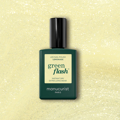 A bottle of Manucurist Lemonade gel polish stands against a yellow glittery background, with a dark green bottle, pale yellow label, and black cap—showcasing the brand&