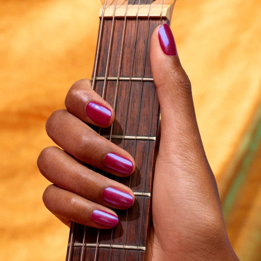 A hand with a glossy magenta manicure using Manucurist Blue Glazed Powder, showing pearly blue shimmer, holds a guitar neck against a warm yellow background.
